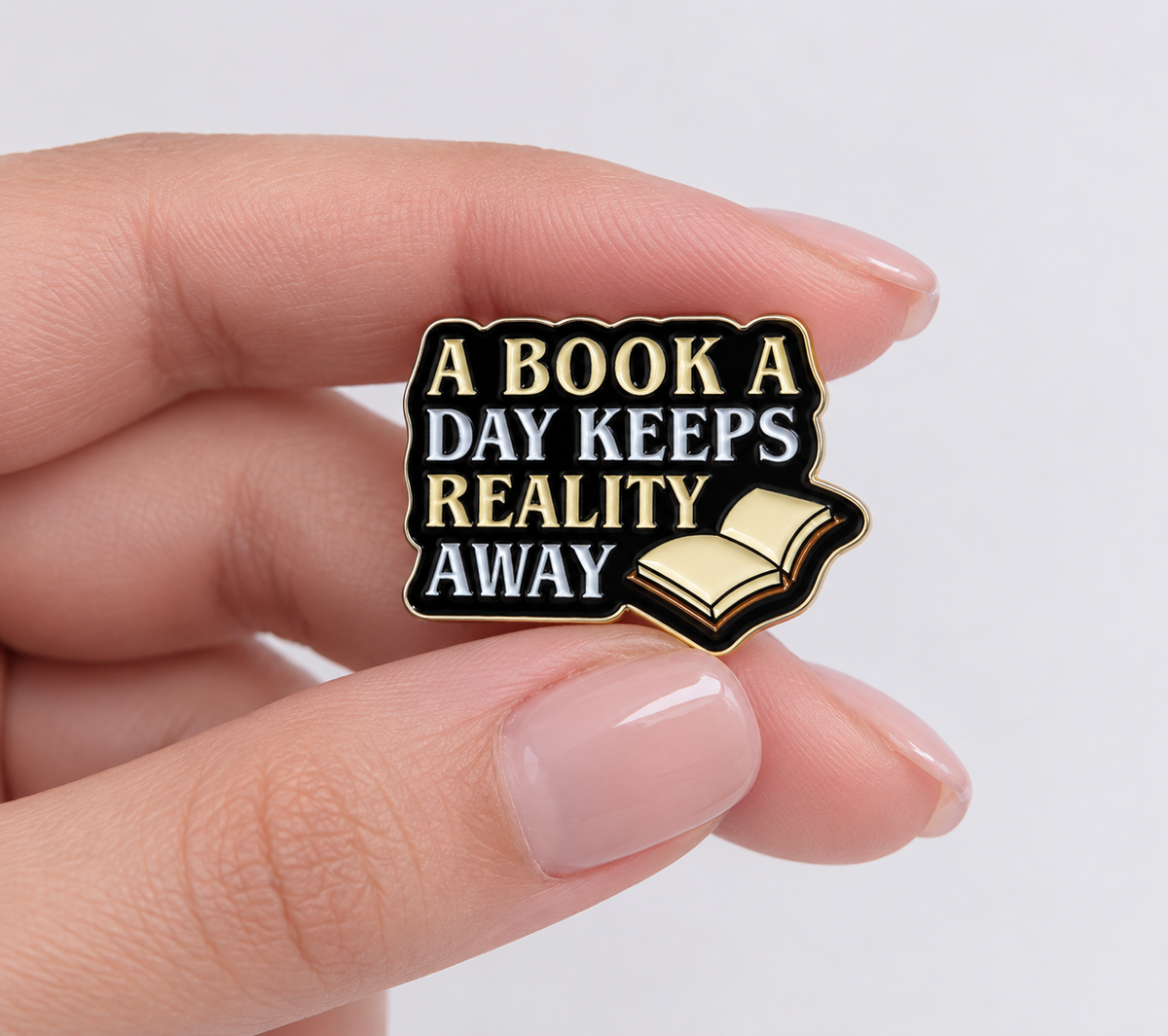 A Book a Day Keeps Reality Away enamel pin held in hand to show its size